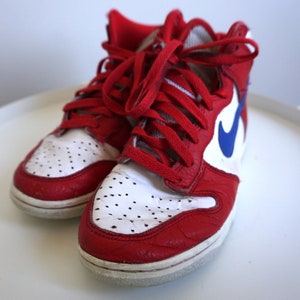 Puede incluir: Par de zapatillas altas rojas y blancas con cordones rojos. Las zapatillas presentan un logotipo azul en forma de &laquo;swoosh&raquo; en el lateral. La puntera tiene peque&ntilde;as perforaciones. Los zapatos est&aacute;n sobre una superficie blanca.