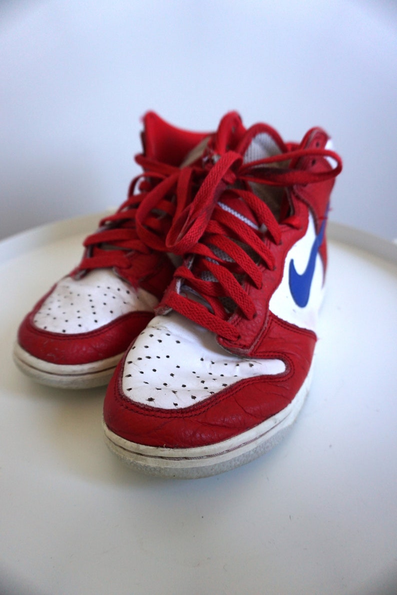 Puede incluir: Par de zapatillas altas rojas y blancas con cordones rojos. Las zapatillas presentan un logotipo azul en forma de &laquo;swoosh&raquo; en el lateral. La puntera tiene peque&ntilde;as perforaciones. Los zapatos est&aacute;n sobre una superficie blanca.