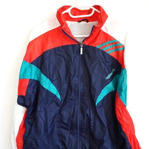 Può includere: Una giacca sportiva a maniche lunghe con zip e design a blocchi di colore in blu navy, rosso acceso e verde acqua. La giacca presenta inserti bianchi sulle spalle, un colletto alto e il logo Adidas ricamato in verde acqua sul petto.