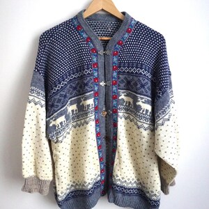 Könnte beinhalten: Ein Vintage-Cardigan im traditionellen nordischen Design. Der Pullover hat ein marineblaues und cremefarbenes Farbschema mit einem Muster aus Schneeflocken und Rentieren. Er hat einen grauen Kragen und Manschetten und wird mit Metallverschlüssen geschlossen. Maße in Zentimetern.