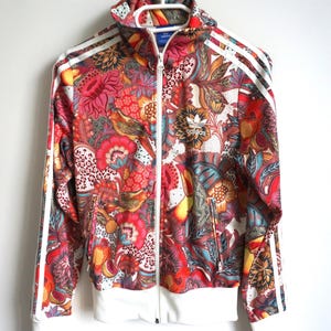 Pode incluir: Um casaco desportivo com fecho de correr apresentando um padrão floral e paisley multicolorido vibrante em tons de vermelho, laranja, amarelo e azul. O casaco inclui um fecho frontal branco, punhos e bainha canelados brancos e riscas brancas.