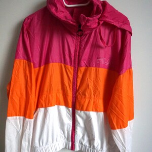 May include: A color-blocked jacket in pink, orange, and white, hanging on a white hanger. The jacket has a hood and a full zipper. The Fila logo is visible on the right chest area.