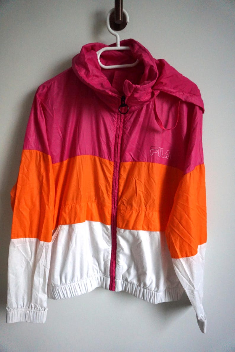 May include: A color-blocked jacket in pink, orange, and white, hanging on a white hanger. The jacket has a hood and a full zipper. The Fila logo is visible on the right chest area.