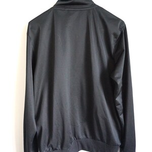 May include: A solid black long-sleeve track jacket featuring a high mock neck collar, a full-length front zipper, and ribbed knit cuffs and hem. The garment is constructed from a smooth, slightly reflective synthetic fabric with a clean design.