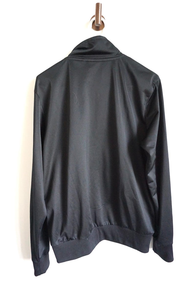 May include: A solid black long-sleeve track jacket featuring a high mock neck collar, a full-length front zipper, and ribbed knit cuffs and hem. The garment is constructed from a smooth, slightly reflective synthetic fabric with a clean design.
