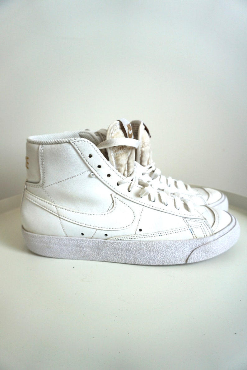 Op de afbeelding: Witte high-top sneakers met een klassiek design. De schoenen hebben het Nike swoosh-logo aan de zijkant en gouden letters op de hiel. De sneakers hebben witte veters en een witte zool.