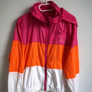 May include: A color-blocked jacket in pink, orange, and white, with a hood and a full-length zipper. The jacket has the brand name "FILA" printed on the chest. The jacket is hanging on a white hanger.