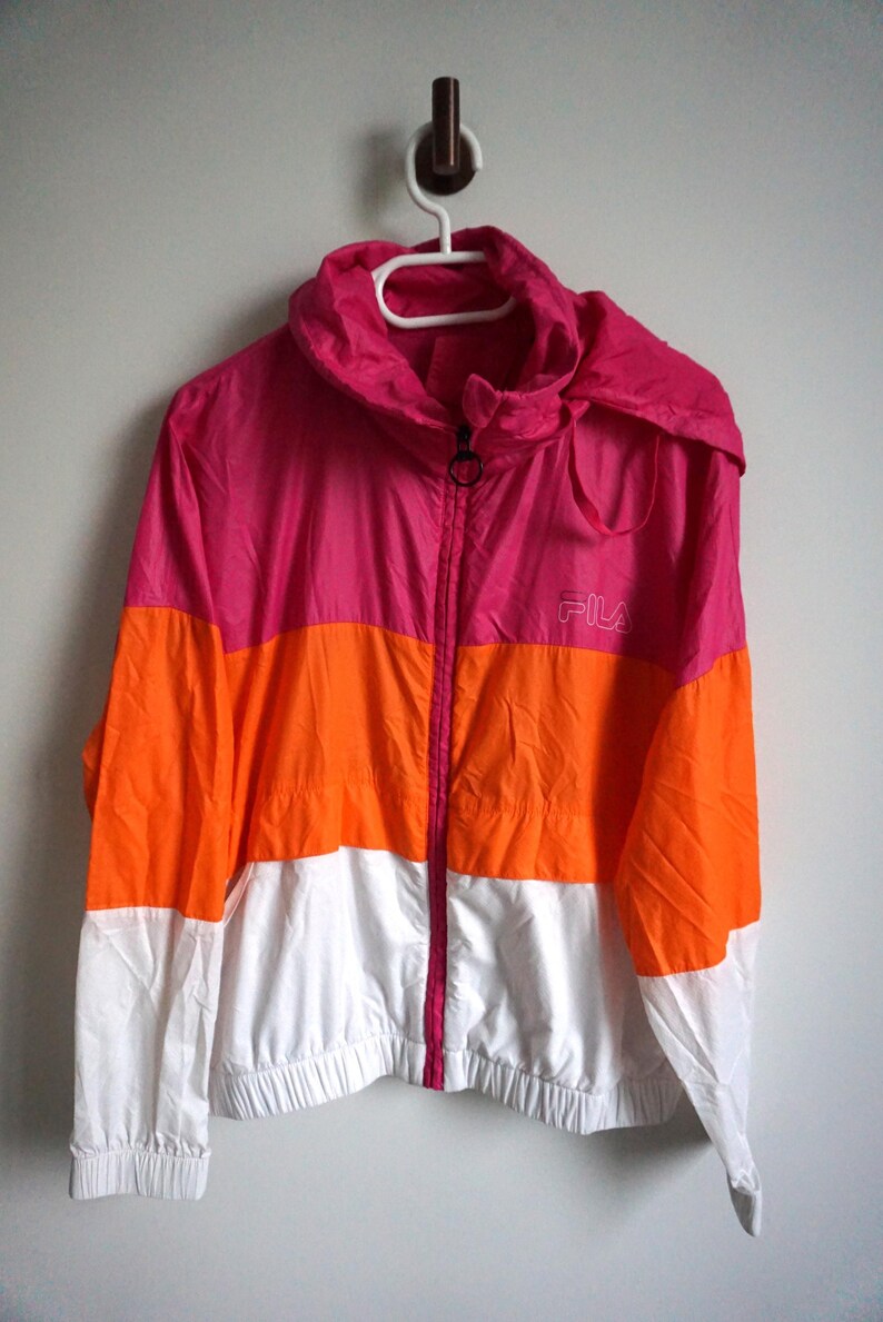 May include: A color-blocked jacket in pink, orange, and white, with a hood and a full-length zipper. The jacket has the brand name "FILA" printed on the chest. The jacket is hanging on a white hanger.