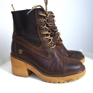 Puede incluir: Botines de cuero marrón con tacón ancho y suela de goma. Las botas tienen un diseño con cordones y ojales metálicos, y un cuello acolchado. El logotipo de Timberland es visible. Altura del tacón: aprox. 7,6 cm.