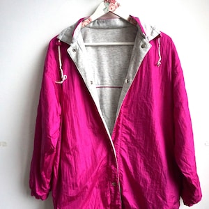 Könnte beinhalten: Eine leuchtend fuchsiafarbene Windjacke mit hellgrau meliertem Innenfutter, einer Kapuze mit weißen Kordelzügen, langen Ärmeln mit elastischen Bündchen und einem Vorderverschluss mit silberfarbenen Metalldruckknöpfen an der Knopfleiste.