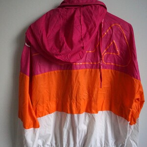 May include: A color-blocked windbreaker jacket in pink, orange, and white. The jacket has a hood and a drawstring. The back of the jacket features a large orange graphic design. The jacket is hanging on a white hook.