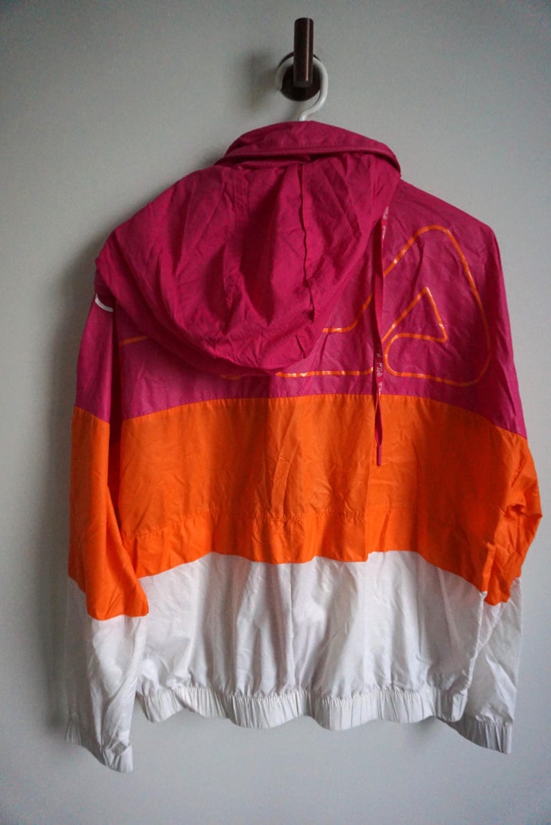 May include: A color-blocked windbreaker jacket in pink, orange, and white. The jacket has a hood and a drawstring. The back of the jacket features a large orange graphic design. The jacket is hanging on a white hook.