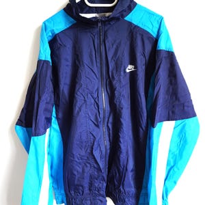 Può includere: Una giacca a vento a maniche lunghe con un design a blocchi di colore in blu navy scuro e blu ciano brillante. La giacca presenta una chiusura frontale con zip, un colletto alto e il logo Nike bianco ricamato sul lato sinistro del petto.