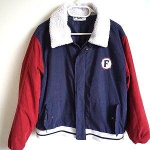 K&ouml;nnte beinhalten: Eine marineblaue und rote Vintage-Jacke mit wei&szlig;em Kunstfellkragen. Die Jacke hat rote &Auml;rmel, einen marineblauen K&ouml;rper und einen wei&szlig;en Streifen am Saum. Ein kreisf&ouml;rmiger Aufn&auml;her mit dem Buchstaben "F" befindet sich auf der Brust.