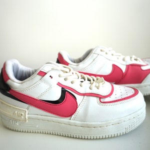 Puede incluir: Un par de zapatillas Nike Air Force 1 blancas y rosas. Las zapatillas presentan una parte superior de cuero blanco con detalles rosas en el logotipo de Swoosh y el tal&oacute;n. La entresuela es blanca y la suela es de color claro.