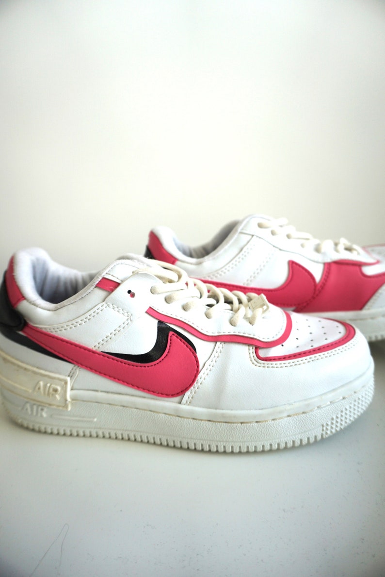 Puede incluir: Un par de zapatillas Nike Air Force 1 blancas y rosas. Las zapatillas presentan una parte superior de cuero blanco con detalles rosas en el logotipo de Swoosh y el tal&oacute;n. La entresuela es blanca y la suela es de color claro.