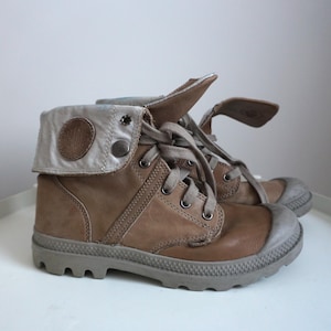 K&ouml;nnte beinhalten: Ein Paar braune Lederstiefel mit grauer Gummisohle. Die Stiefel haben einen umklappbaren Schaft mit silberfarbenem Akzent und Knopfverschluss. Die Schn&uuml;rsenkel sind hellbraun. Die Stiefel sind etwa 15 cm hoch.