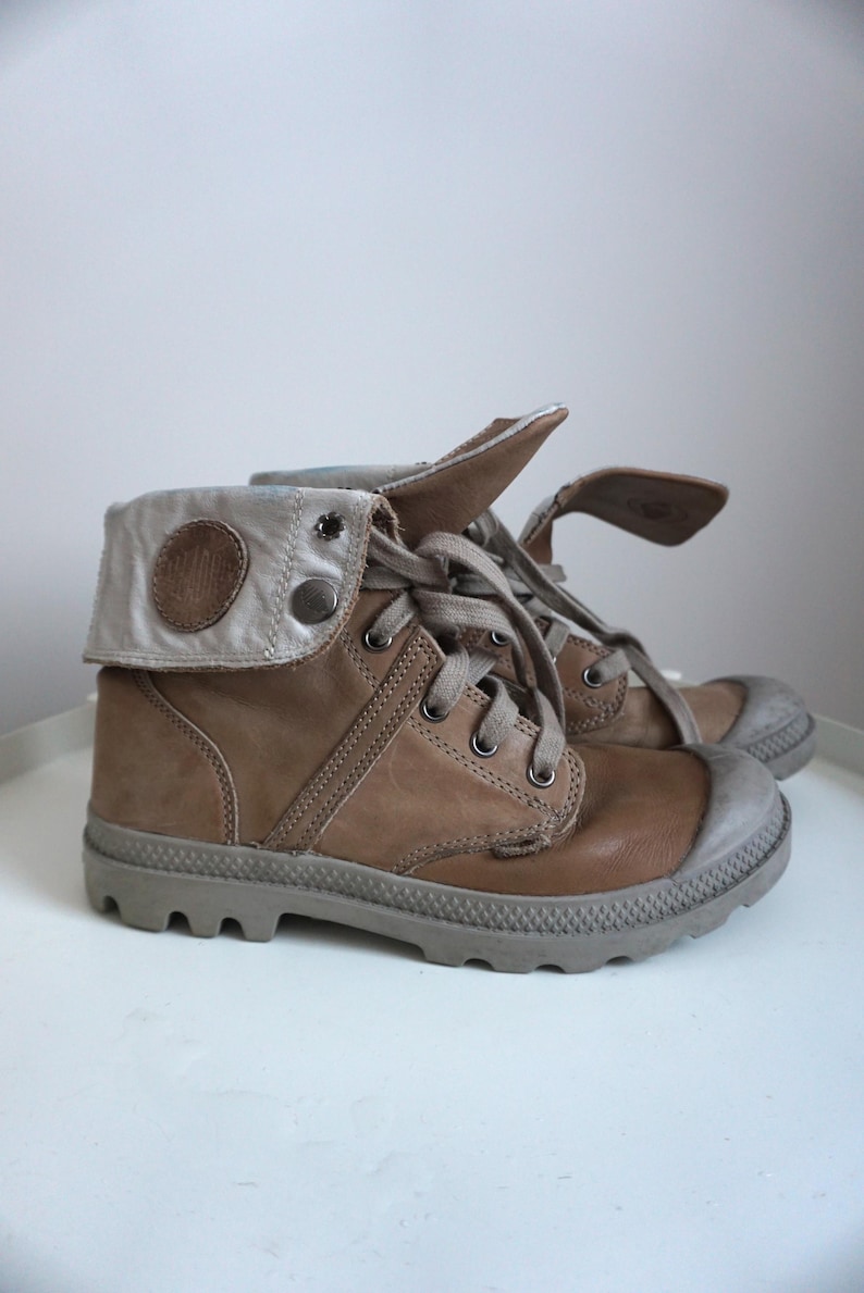 K&ouml;nnte beinhalten: Ein Paar braune Lederstiefel mit grauer Gummisohle. Die Stiefel haben einen umklappbaren Schaft mit silberfarbenem Akzent und Knopfverschluss. Die Schn&uuml;rsenkel sind hellbraun. Die Stiefel sind etwa 15 cm hoch.