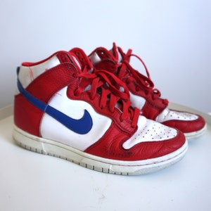 Puede incluir: Par de zapatillas altas rojas, blancas y azules. Las zapatillas presentan una parte superior de cuero rojo con paneles blancos y un logotipo azul de la marca. Los zapatos tienen cordones rojos y una suela blanca.