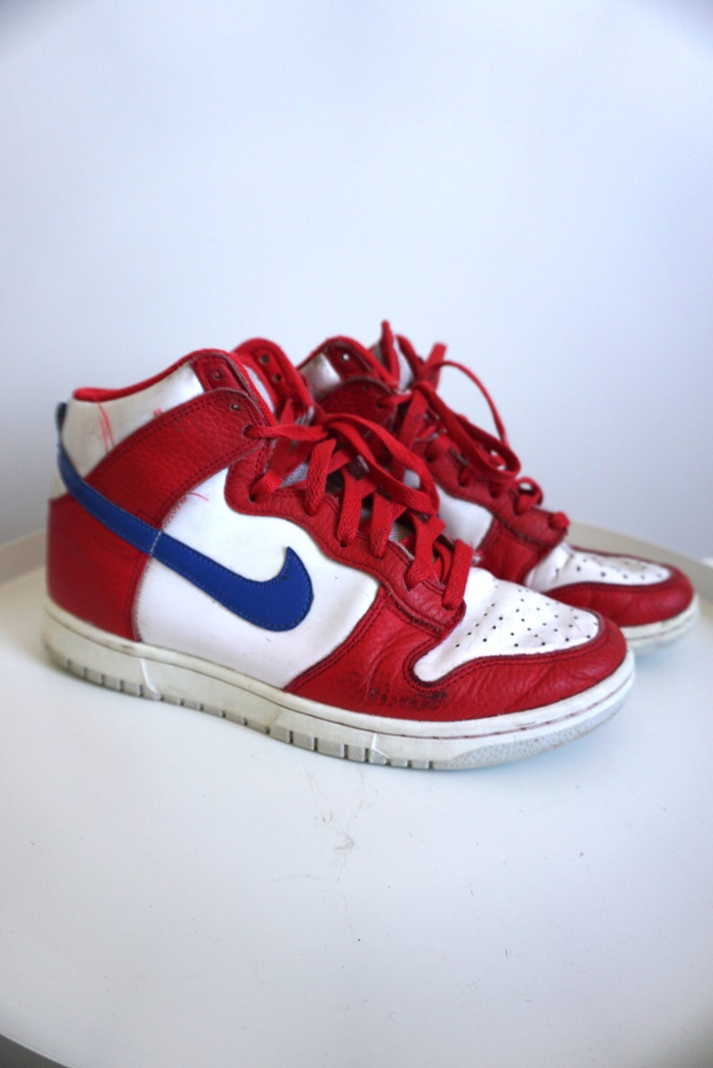 Puede incluir: Par de zapatillas altas rojas, blancas y azules. Las zapatillas presentan una parte superior de cuero rojo con paneles blancos y un logotipo azul de la marca. Los zapatos tienen cordones rojos y una suela blanca.