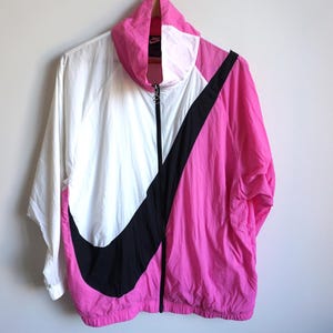 Könnte beinhalten: Eine weiße, rosa und schwarze Nike Windjacke mit großem schwarzem Swoosh-Logo. Die Jacke hat einen durchgehenden Reißverschluss und einen hohen Kragen. Die Jacke hängt an einer Wand.