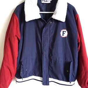 K&ouml;nnte beinhalten: Eine marineblaue Vintage-Fila-Jacke mit roten &Auml;rmeln und einem flauschigen wei&szlig;en Kragen. Die Jacke hat einen wei&szlig;-marinefarbenen gestreiften Saum und einen runden Aufn&auml;her mit dem Buchstaben "F". Das Fila-Logo ist am Kragen sichtbar.
