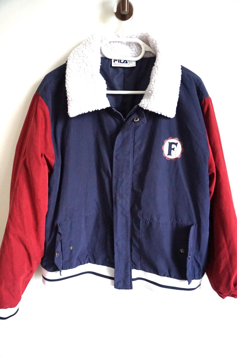 K&ouml;nnte beinhalten: Eine marineblaue Vintage-Fila-Jacke mit roten &Auml;rmeln und einem flauschigen wei&szlig;en Kragen. Die Jacke hat einen wei&szlig;-marinefarbenen gestreiften Saum und einen runden Aufn&auml;her mit dem Buchstaben "F". Das Fila-Logo ist am Kragen sichtbar.