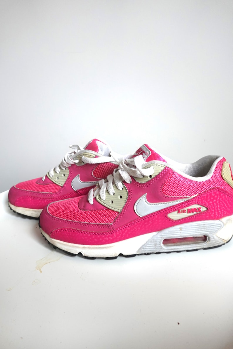 K&ouml;nnte beinhalten: Paar leuchtend pinkfarbene Air Max 90 Sneaker. Die Schuhe haben wei&szlig;e Schn&uuml;rsenkel, einen wei&szlig;en Nike-Swoosh und eine wei&szlig;e und graue Sohle. Die Worte "AIR MAX" sind auf der Seite des Schuhs zu sehen.