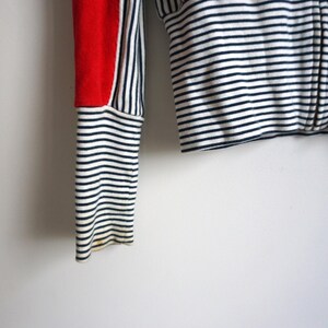 May include: Close-up of a red, white, and navy blue striped long-sleeved top. The top features a red panel on the sleeve and a horizontal striped pattern on the body and sleeves. The sleeve cuff is a navy and white striped pattern.