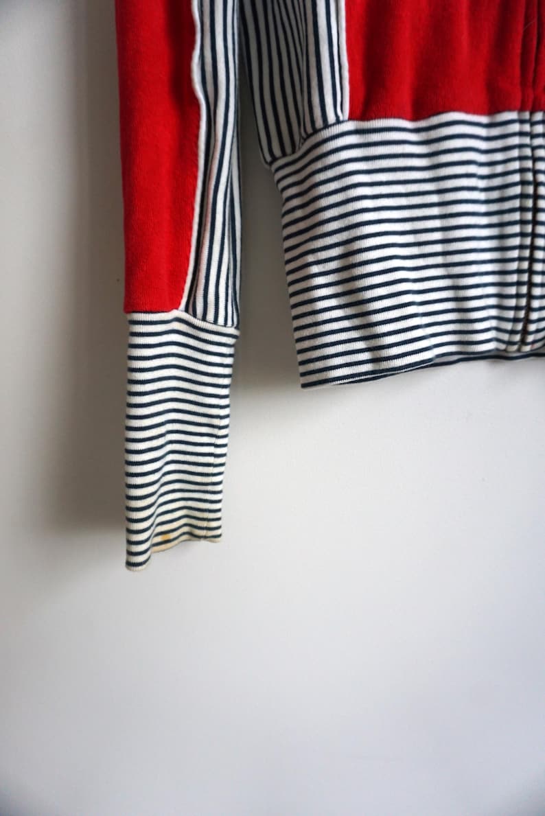 May include: Close-up of a red, white, and navy blue striped long-sleeved top. The top features a red panel on the sleeve and a horizontal striped pattern on the body and sleeves. The sleeve cuff is a navy and white striped pattern.