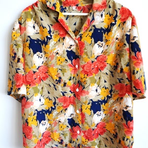Può includere: Una camicia a maniche corte con abbottonatura frontale caratterizzata da un vivace motivo floreale con fiori rossi, gialli e bianchi su sfondo blu scuro e verde oliva. La camicia presenta un colletto classico, orlo dritto e bottoni bianchi.