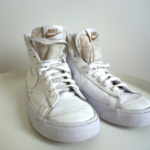 Op de afbeelding: Een paar witte Nike Blazer high-top sneakers. De schoenen hebben witte veters, een witte zool en een gouden Nike-logo op de tong. De sneakers zijn gemaakt van leer en hebben een klassiek ontwerp.