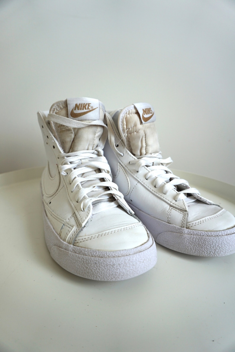 Op de afbeelding: Een paar witte Nike Blazer high-top sneakers. De schoenen hebben witte veters, een witte zool en een gouden Nike-logo op de tong. De sneakers zijn gemaakt van leer en hebben een klassiek ontwerp.