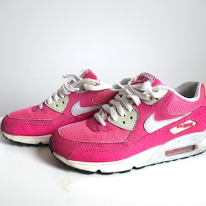 K&ouml;nnte beinhalten: Paar leuchtend pinkfarbene Nike Air Max 90 Sneaker. Die Schuhe haben wei&szlig;e Schn&uuml;rsenkel, einen wei&szlig;en Nike Swoosh und eine wei&szlig;e Zwischensohle. Das Air Max-Logo ist sichtbar. Die Sneaker stehen auf einer wei&szlig;en Oberfl&auml;che.
