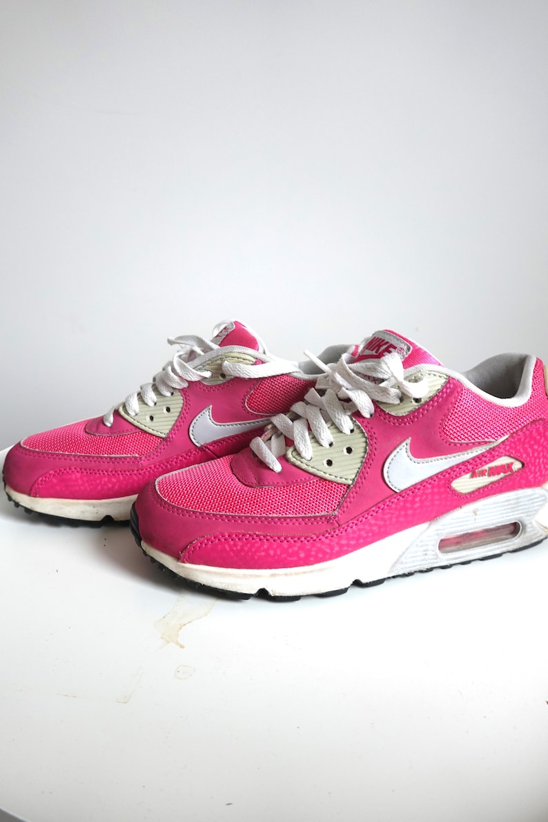 K&ouml;nnte beinhalten: Paar leuchtend pinkfarbene Nike Air Max 90 Sneaker. Die Schuhe haben wei&szlig;e Schn&uuml;rsenkel, einen wei&szlig;en Nike Swoosh und eine wei&szlig;e Zwischensohle. Das Air Max-Logo ist sichtbar. Die Sneaker stehen auf einer wei&szlig;en Oberfl&auml;che.