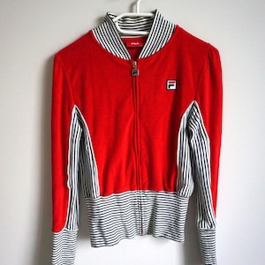 May include: A red zip-up jacket with black and white striped accents. The jacket features a stand-up collar and the Fila logo on the chest. The sleeves and lower hem have black and white stripes. The jacket is hanging on a white hanger.