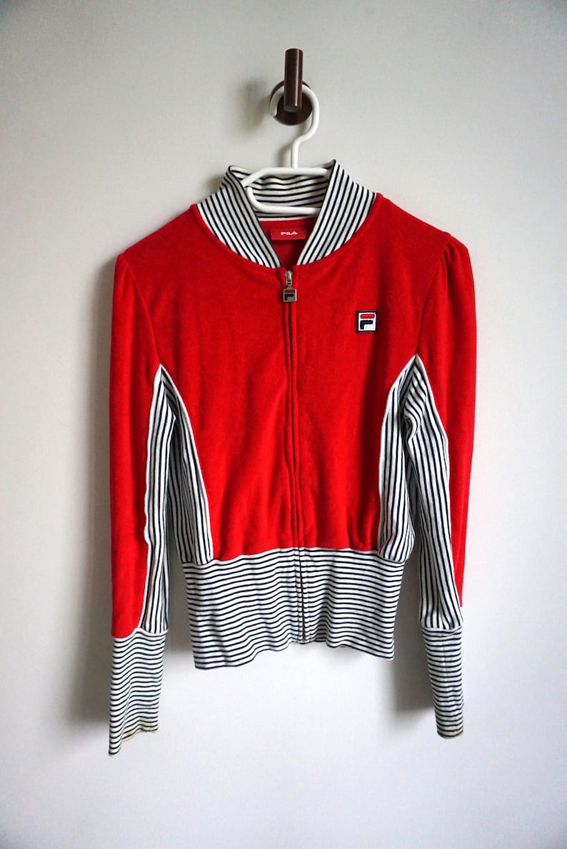 May include: A red zip-up jacket with black and white striped accents. The jacket features a stand-up collar and the Fila logo on the chest. The sleeves and lower hem have black and white stripes. The jacket is hanging on a white hanger.