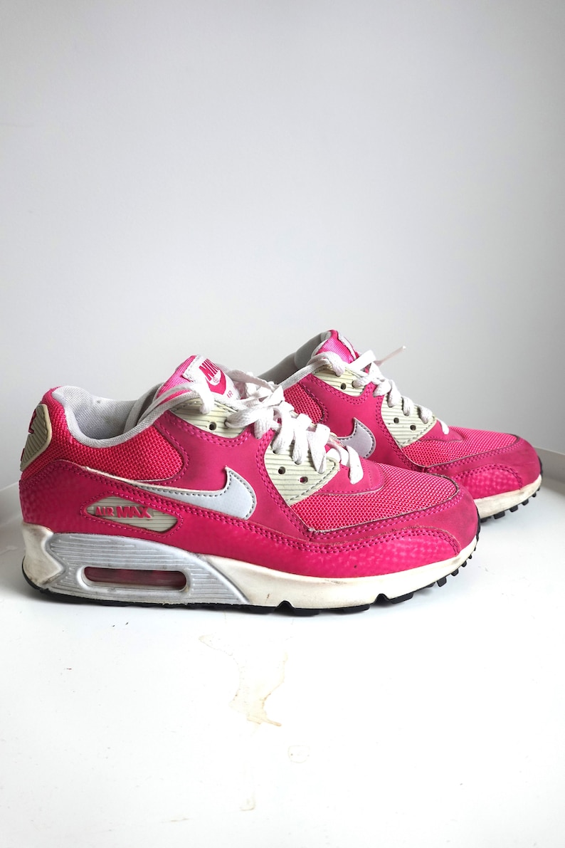 K&ouml;nnte beinhalten: Paar leuchtend pinkfarbene Nike Air Max 90 Sneaker. Die Schuhe haben wei&szlig;e Schn&uuml;rsenkel, einen wei&szlig;en Swoosh und eine wei&szlig;-graue Sohle. Das Air Max-Logo ist seitlich sichtbar. Die Sneaker stehen auf einer wei&szlig;en Oberfl&auml;che.
