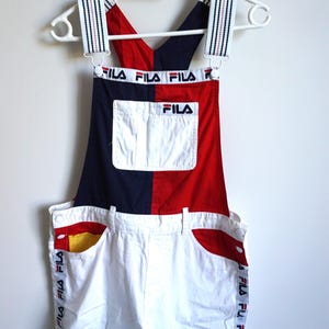 Puede incluir: Mono Fila vintage en blanco, azul marino y rojo. El mono con peto presenta un dise&ntilde;o de bloques de color con el logotipo de Fila en el bolsillo del peto y a lo largo del lateral del pantal&oacute;n corto. Las correas est&aacute;n rayadas con el logotipo de Fila.