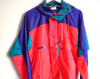 May include: A vintage windbreaker jacket in a vibrant color block design. The jacket features a hood, pockets, and a drawstring. The color scheme includes bright coral, teal, and purple. The jacket is hanging on a white wall.