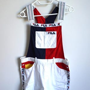 Puede incluir: Un conjunto de peto corto Fila vintage. La parte superior es roja y azul marino, con el logo de Fila. Los pantalones cortos blancos tienen detalles de bolsillo rojos y amarillos y la marca Fila en el lateral. Las correas son rayadas.