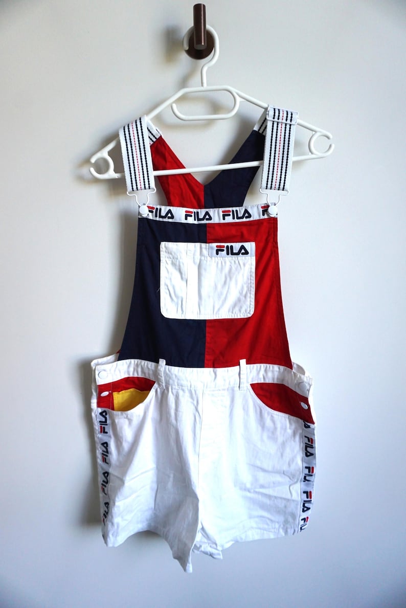 Puede incluir: Un conjunto de peto corto Fila vintage. La parte superior es roja y azul marino, con el logo de Fila. Los pantalones cortos blancos tienen detalles de bolsillo rojos y amarillos y la marca Fila en el lateral. Las correas son rayadas.