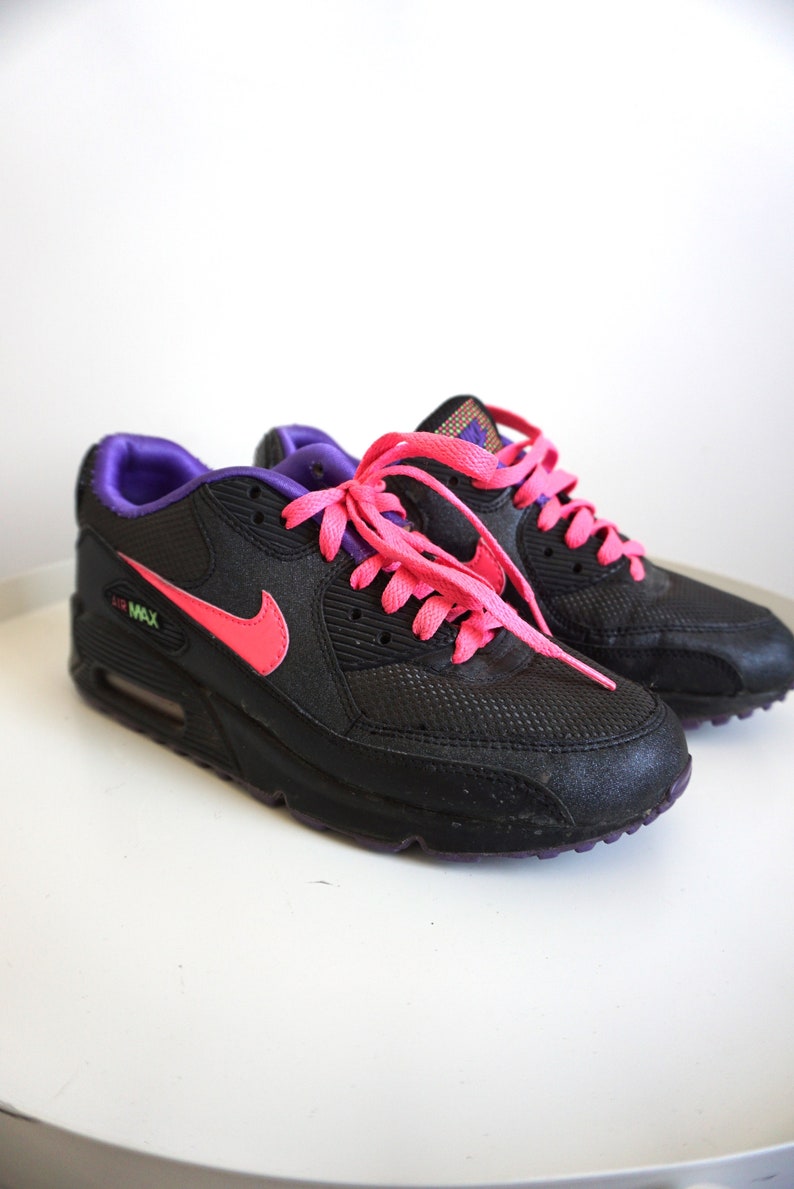 May include: Black Nike Air Max 90 sneakers with pink laces and a purple trim. The shoes have a mesh and leather upper and a white midsole.