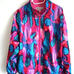 Könnte beinhalten: Eine langärmelige Jacke mit Reißverschluss und einem lebhaften abstrakten Muster in den Farben Pink, leuchtendem Blau und dunklem Türkis. Die Jacke hat einen hohen Kragen, einen durchgehenden pinken Reißverschluss sowie elastische Bündchen und Saum. Das Etikett zeigt CHAVER.