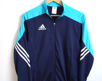 Könnte beinhalten: Marineblaue und türkisfarbene Adidas-Trainingsjacke mit durchgehendem Reißverschluss. Die Jacke hat das Adidas-Logo auf der Brust und drei weiße Streifen auf den Ärmeln. Die Jacke hängt an einem Holzbügel.