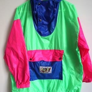 Può includere: Una giacca a vento verde neon con maniche rosa acceso, cappuccio blu e una tasca frontale blu con patta. La tasca presenta una piccola toppa rettangolare con un'illustrazione grafica di figure. La giacca ha un orlo con coulisse.