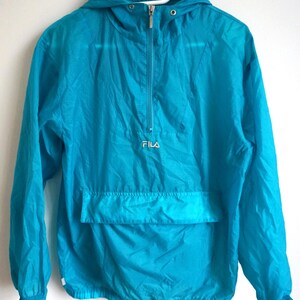 Pode incluir: Um blus&atilde;o corta-vento turquesa brilhante com capuz, mangas compridas com punhos el&aacute;sticos e fecho de correr frontal at&eacute; meio. A pe&ccedil;a inclui um bolso frontal horizontal grande e o nome da marca FILA bordado a branco no peito.
