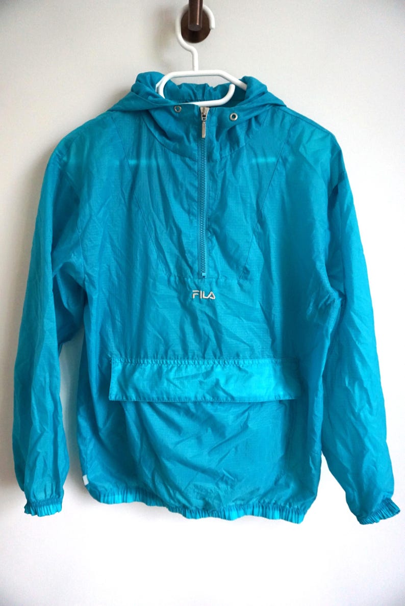 Pode incluir: Um blus&atilde;o corta-vento turquesa brilhante com capuz, mangas compridas com punhos el&aacute;sticos e fecho de correr frontal at&eacute; meio. A pe&ccedil;a inclui um bolso frontal horizontal grande e o nome da marca FILA bordado a branco no peito.