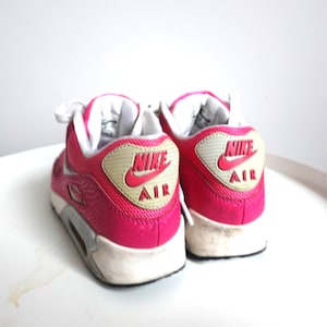 K&ouml;nnte beinhalten: Paar leuchtend pinkfarbene Nike Air Max 90 Sneaker. Die Schuhe haben wei&szlig;e Schn&uuml;rsenkel, eine wei&szlig;e Sohle und eine grau-wei&szlig;e Zwischensohle. Das Nike-Logo und "AIR" sind rot auf einem beigefarbenen Patch an der Ferse. Auf einer wei&szlig;en Oberfl&auml;che.