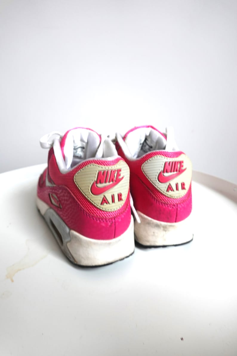 K&ouml;nnte beinhalten: Paar leuchtend pinkfarbene Nike Air Max 90 Sneaker. Die Schuhe haben wei&szlig;e Schn&uuml;rsenkel, eine wei&szlig;e Sohle und eine grau-wei&szlig;e Zwischensohle. Das Nike-Logo und "AIR" sind rot auf einem beigefarbenen Patch an der Ferse. Auf einer wei&szlig;en Oberfl&auml;che.